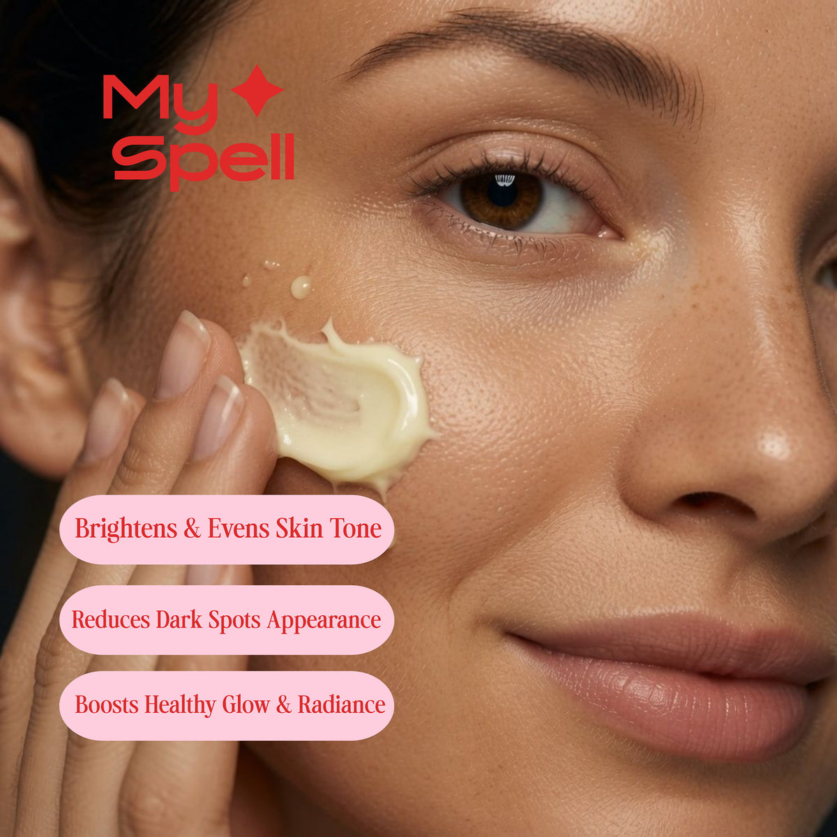 Close-up of a woman applying a cream-colored skincare product, My Spell Vitamin C Serum to her cheek, with the My Spell brand logo and three pink text badges describing skin benefits.