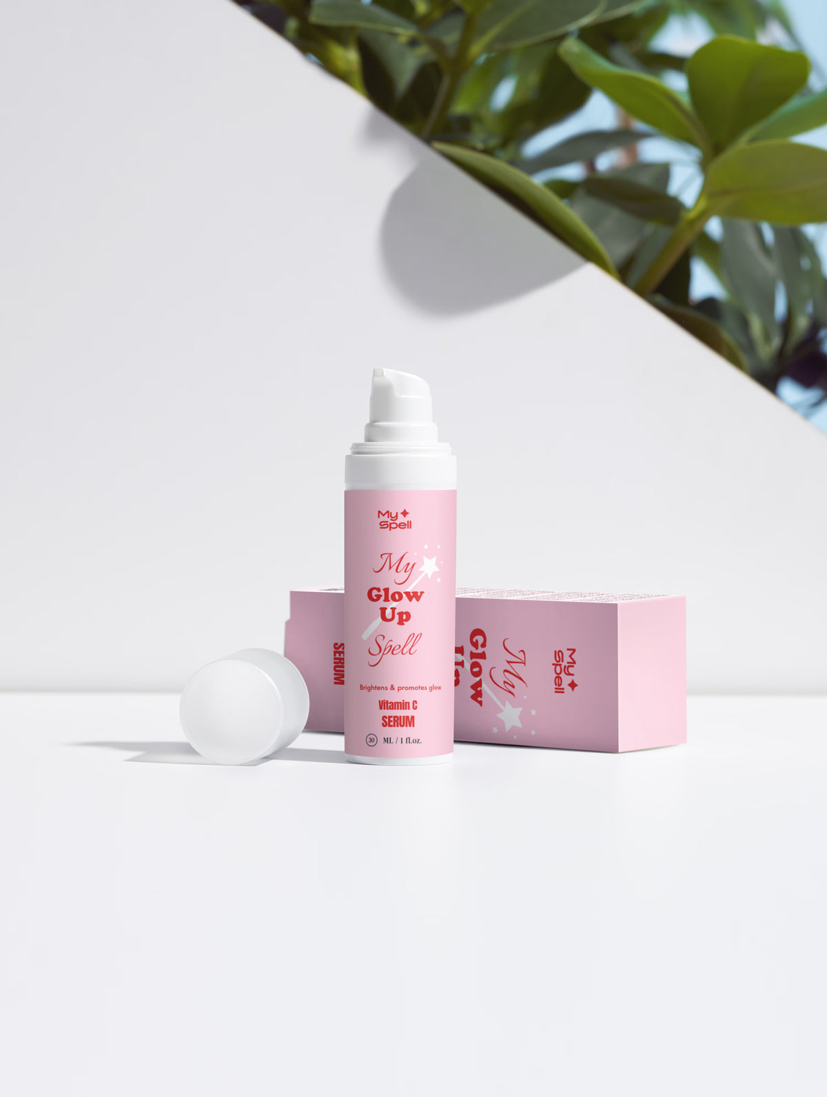 A pink cosmetic Vitamin C serum bottle labeled 'My Glow Up Spell' stands on a white surface with its cap placed beside it, alongside a matching pink box. The scene is styled with a diagonal white line and green leaves in the upper right corner.