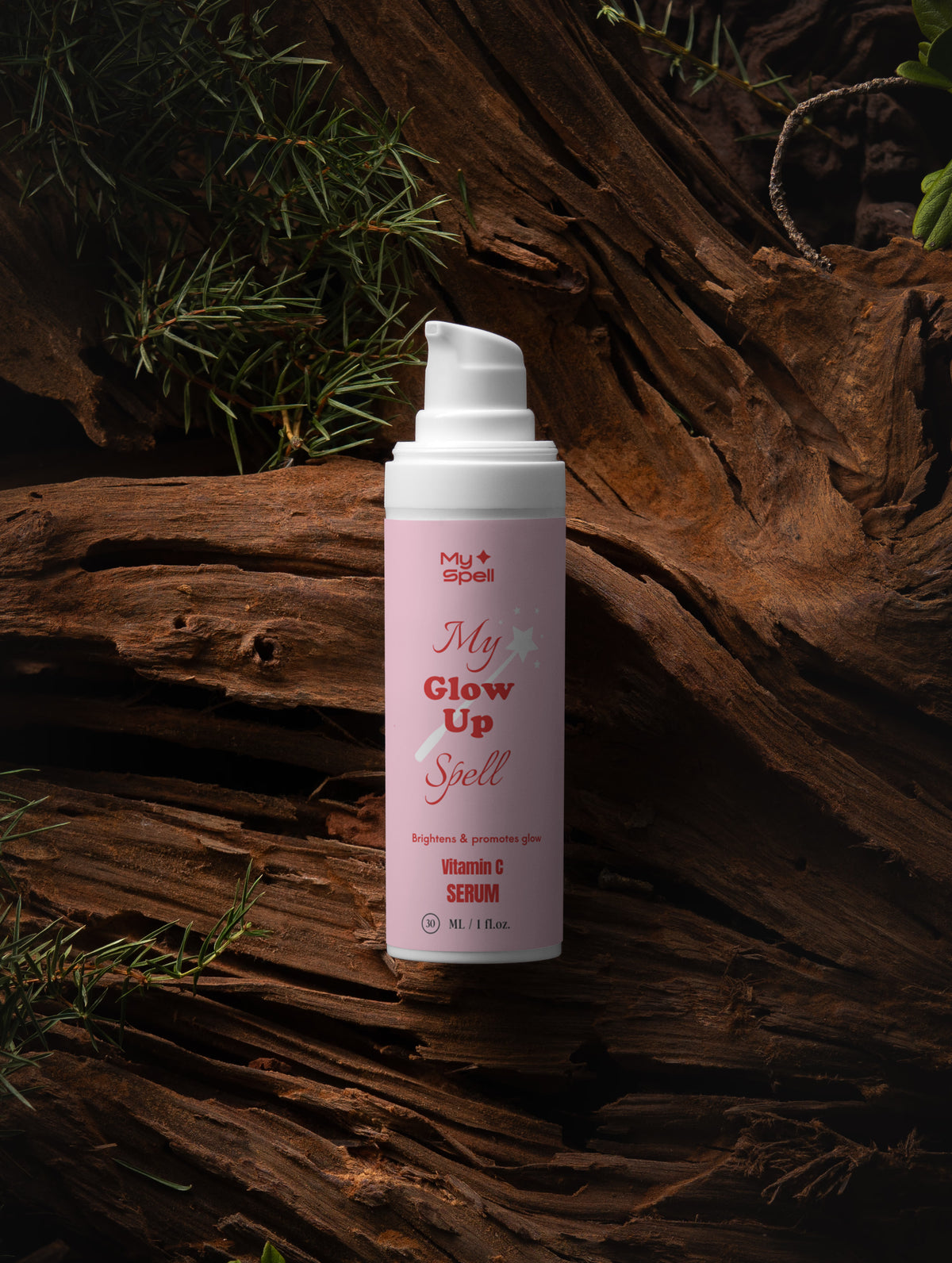 A pump bottle of My Spell, My Glow Up Spell Vitamin C Serum, featuring a light pink label and white pump, set against a dark, rustic background of weathered wood and pine needles.